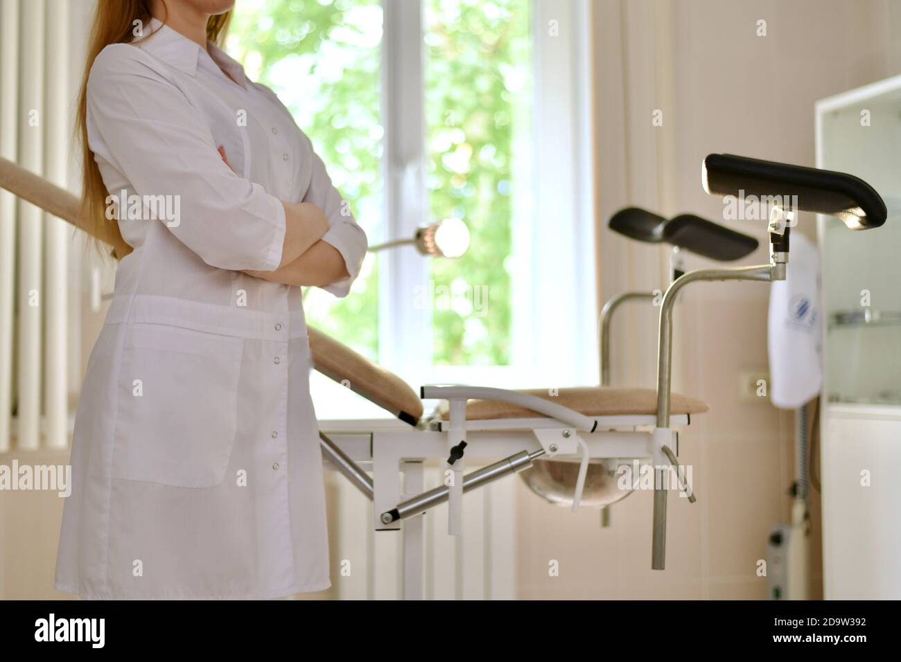 A doctor in white uniform with folded hands. Stands in profile in front of a gynecological chair with footrests.  Stock Photo