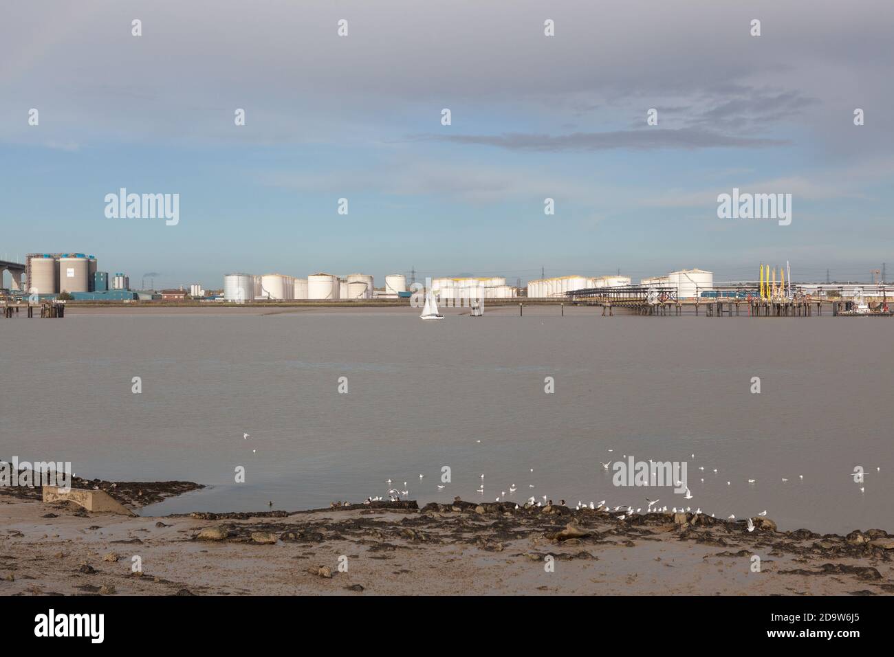 Small white sail boat in the Thames Estuary sails past Oil Storage Depot in Thurrock, Essex, UK. Stock Photo