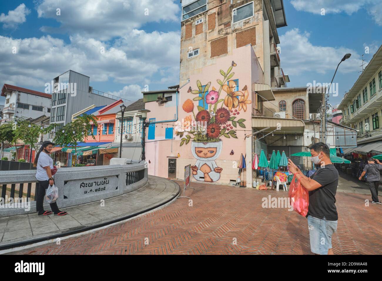 A couple takes photos at a mural by Thai artist Alex Face featuring  his three-eyed character Mardi meditating; Phahurat / Chinatown Bangkok, Thailand Stock Photo