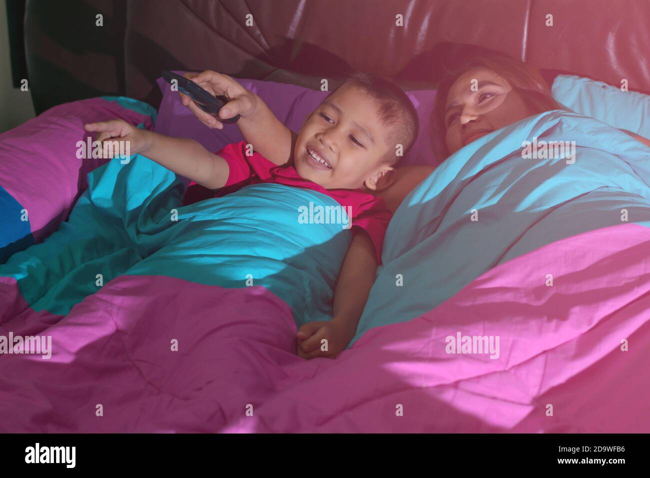 Mother and son sleeps on the bed and holds the TV remote and smiles