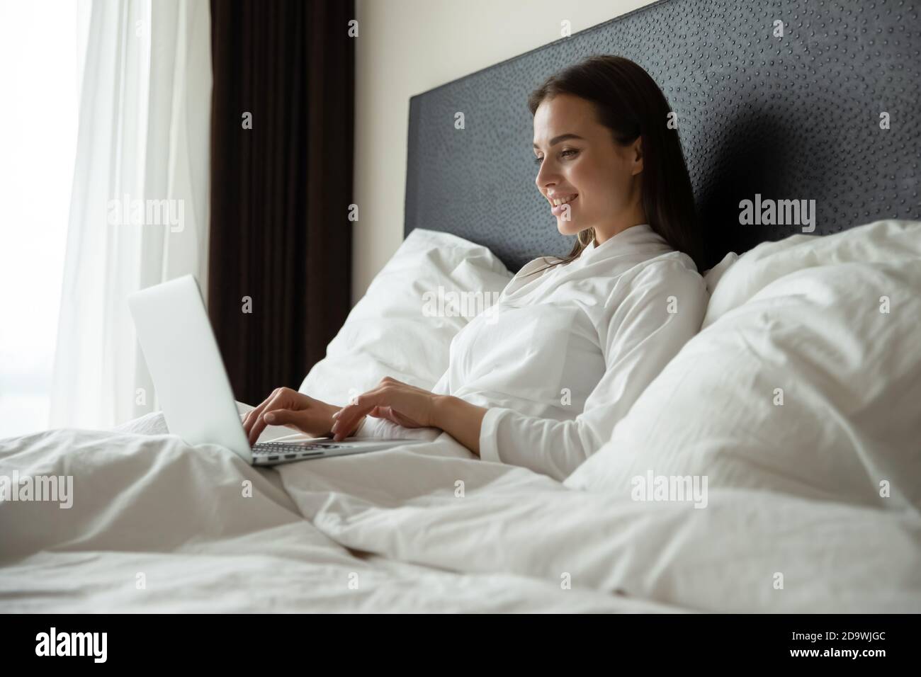 Happy young female remote employee using laptop in comfortable bed