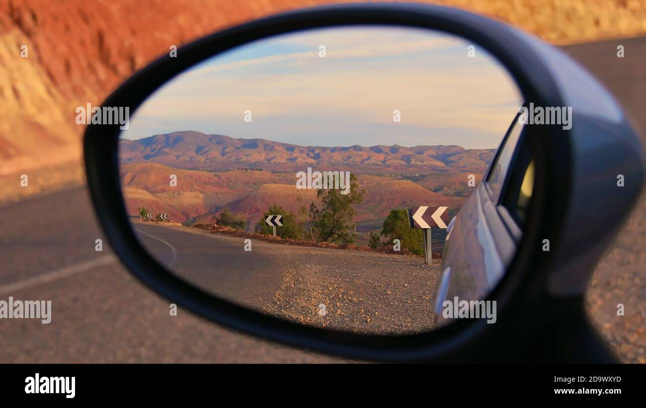 Beautifully red colored foothills of the stunning Atlas Mountains in the evening sun with paved street and road signs viewed in the side mirror of car. Stock Photo