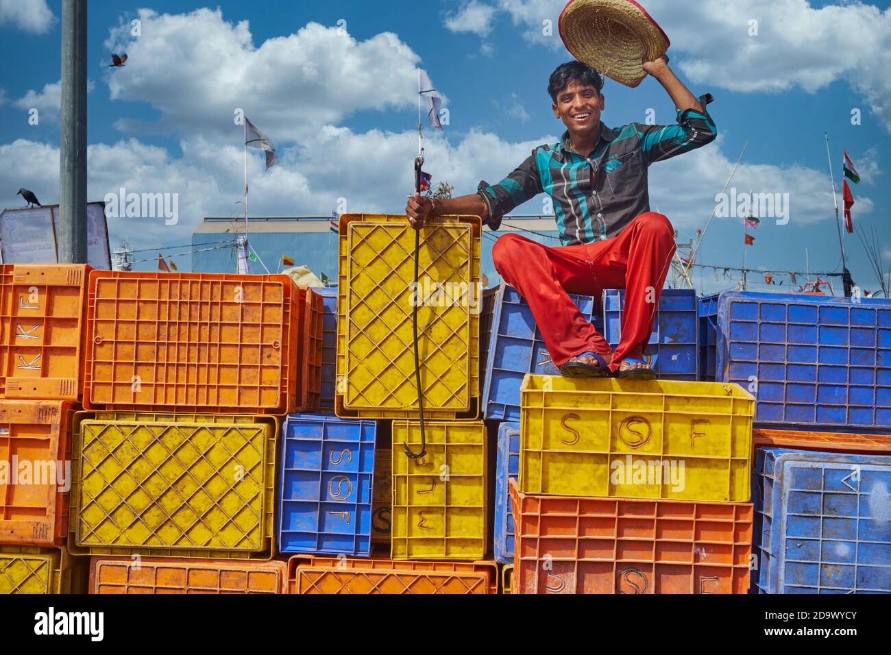 A young Indian man sitting on colorful plastic crates in the Old Port of Mangalore, Karnataka, India, lifts his hat in a cheerful greeting Stock Photo