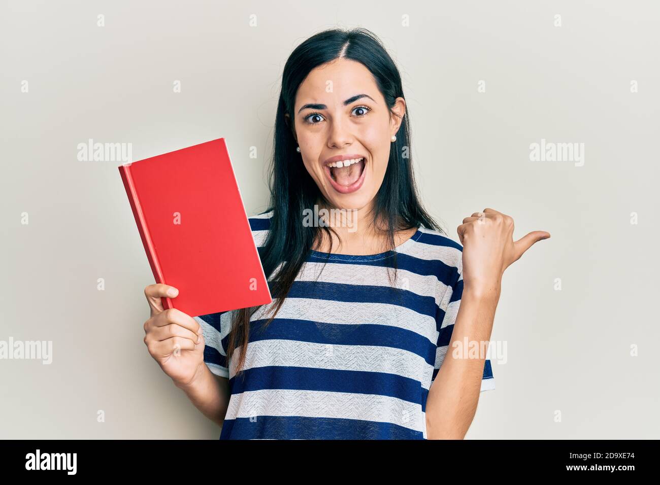 Beautiful young woman reading book pointing thumb up to the side ...