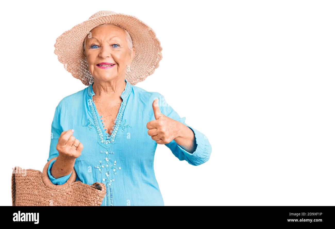 Senior beautiful woman with blue eyes and grey hair wearing fashion dress and hat holding summer wicker handbag smiling happy and positive, thumb up d Stock Photo