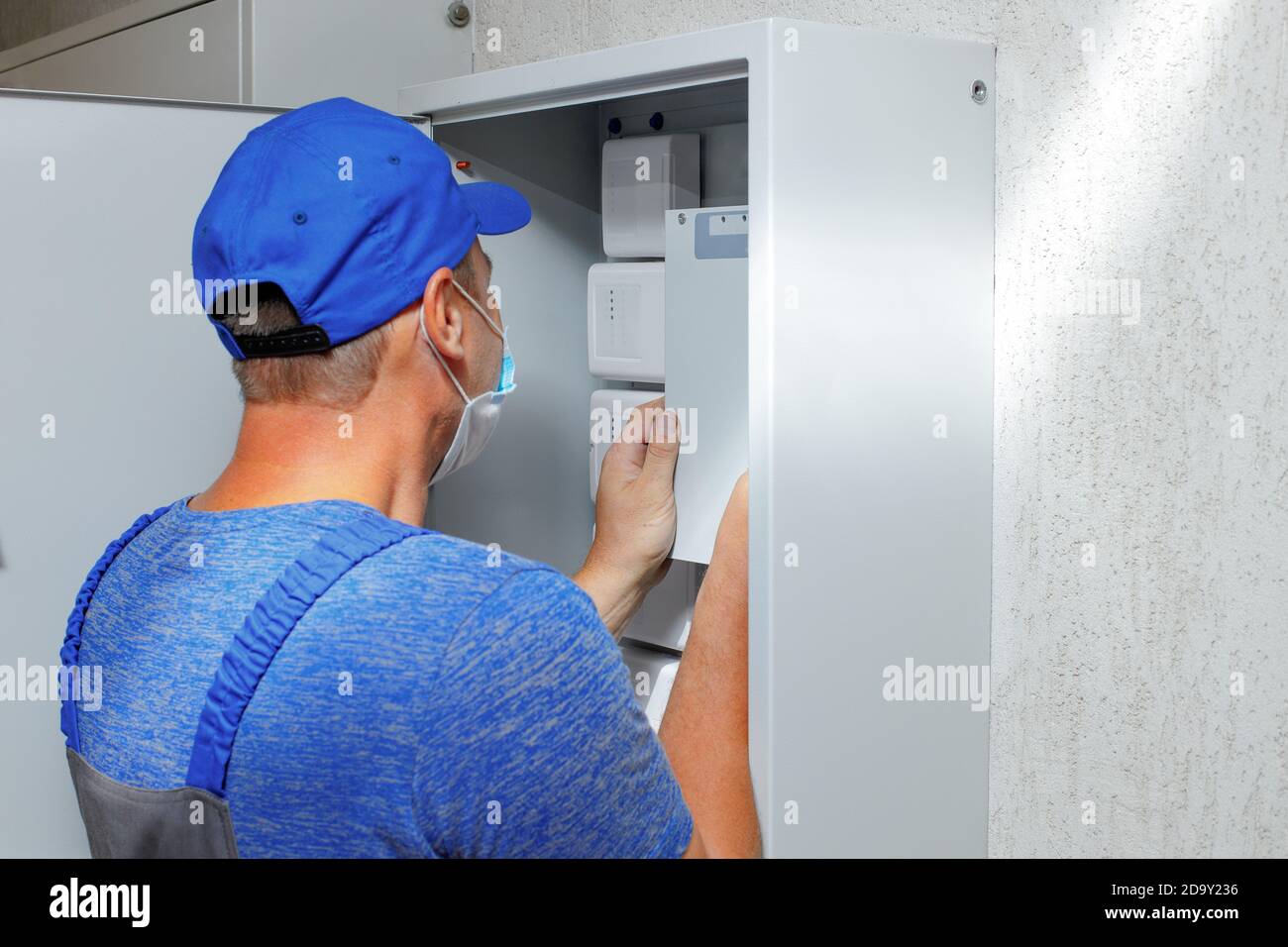 An electrician in overalls and a baseball cap examines a Cabinet with sensors and equipment.  Stock Photo