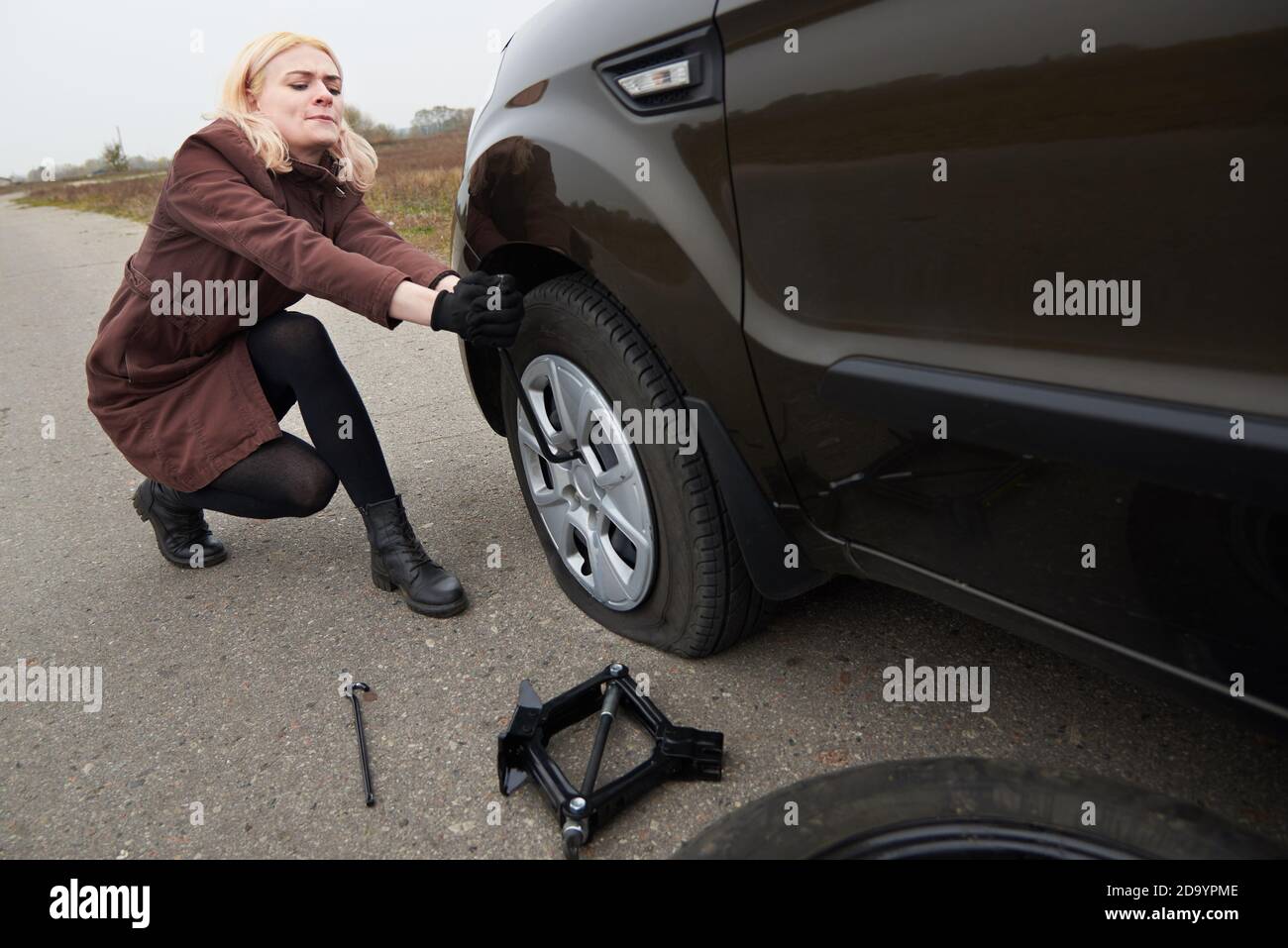 A Young Blonde Woman Removes The Wheel With A Key Near Her Car With A