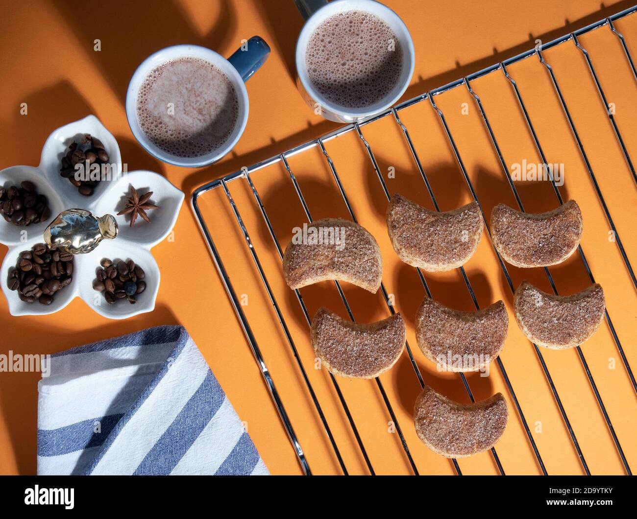Two mugs of cacao, semilunar candied cookies on a metal grate, striped towel and spices. Top view with hard shadows Stock Photo