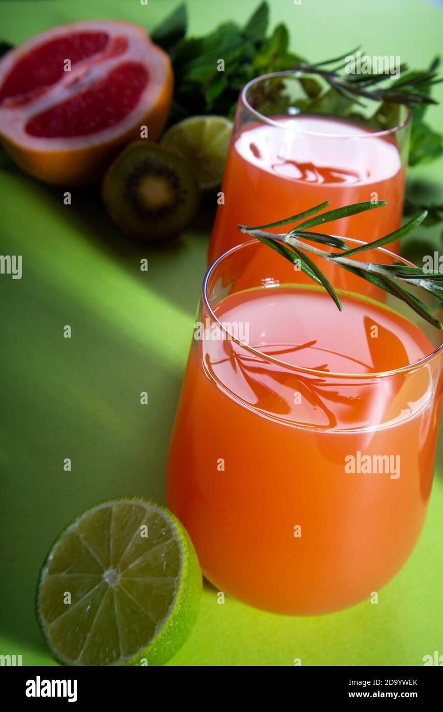 Composition of glasses with orange juice, cut red orange, lime, kiwi, rosemary and mint leaves on a light green background Stock Photo