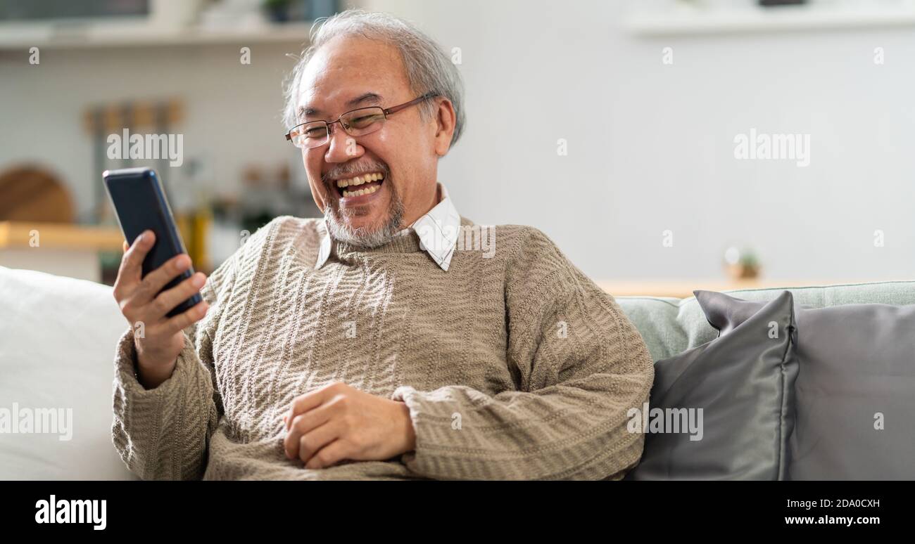 Panoramic Happy retirement elderly man sitting on sofa at living room use cellphone to make video call to his family with laugh and smile. Communicati Stock Photo