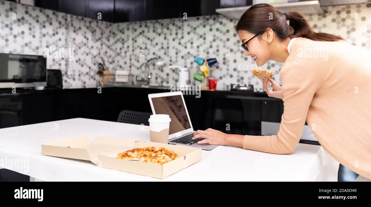 Panoramic Asian woman work from home in the kitchen and eating delivery pizza take away food and take out coffee while city lockdown from coronavirus Stock Photo
