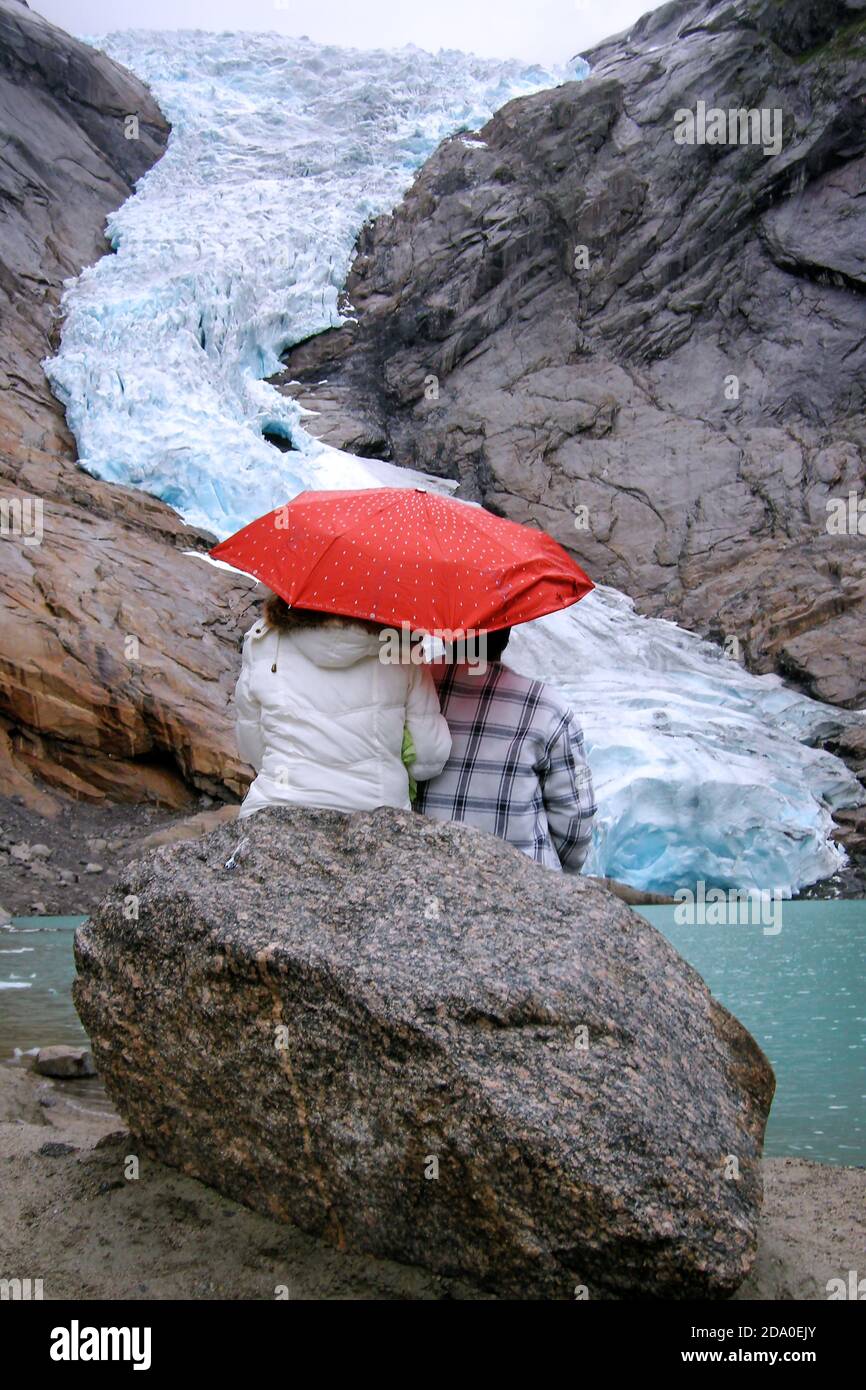 Briksdal Glacier (Briksdalbreen), Norway. Couple with an umbrella sitting on a rock looking at Briskdal Glacier. View of the glacier 10 years ago. Stock Photo
