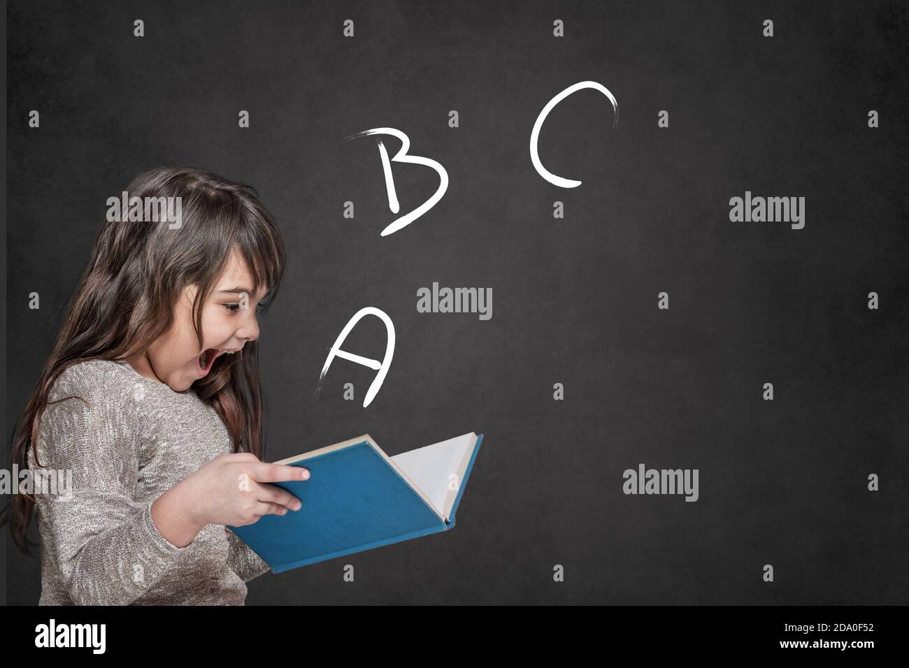 Cute tanned long haired  little girl is surprised at the letters ABC coming out of an open book she is holding in her hands. Stock Photo