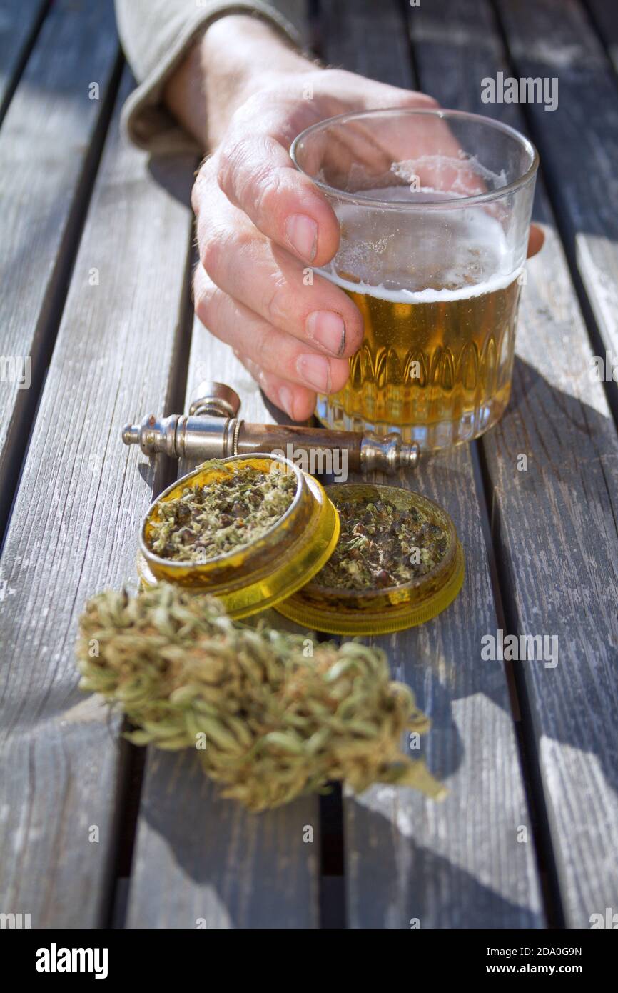 Close up of wood table with marijuana grinder, marijuana bud, a smoking pipe and male hand holding a glass of beer in the background. Stock Photo