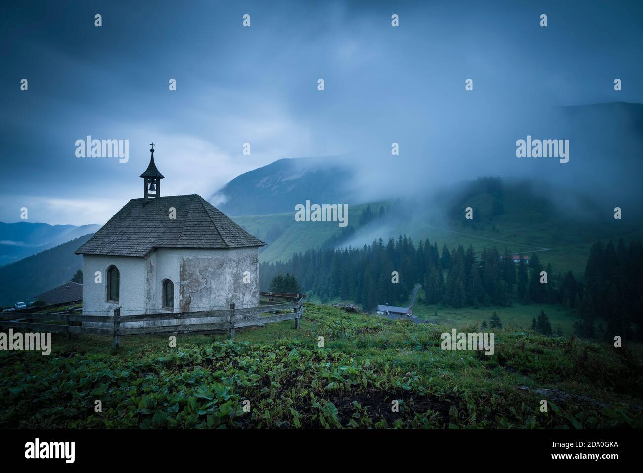 White, shingle-roofed chapel on the Ackernalm alp meadows in front of a mountain panorama with overcast sky and waves of mist, Tyrol, Austria Stock Photo