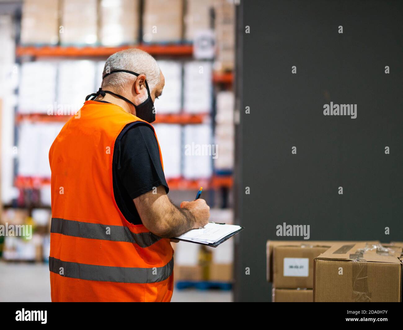 Back view adult worker wearing uniform and protective face mask writing on clipboard while working in spacious storehouse Stock Photo