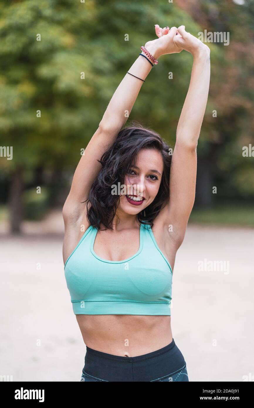 Athletic female in sports bra standing in park and stretching raised arms above head while preparing for training and looking at camera Stock Photo
