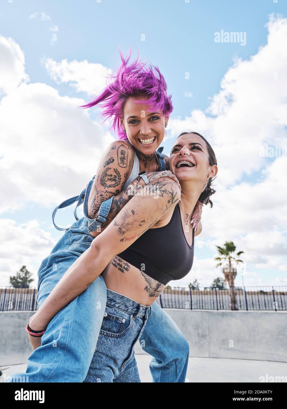 Low angle of delighted Woman with pink hair piggybacking cheerful female friend with tattoos while having fun together at weekend Stock Photo
