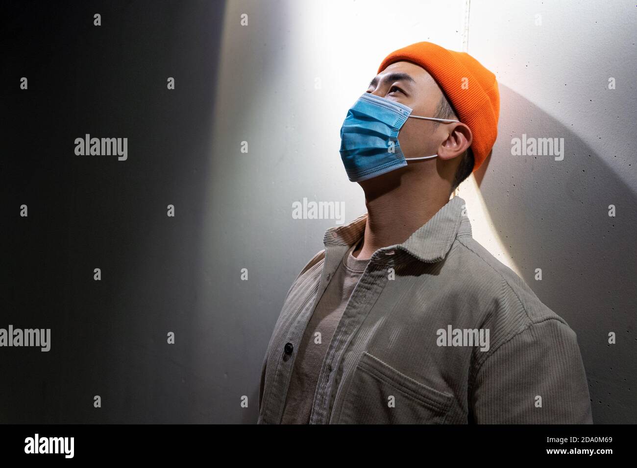Concentrated Asian male in casual wear and protective face mask standing calmly in blurred gray room and looking away Stock Photo