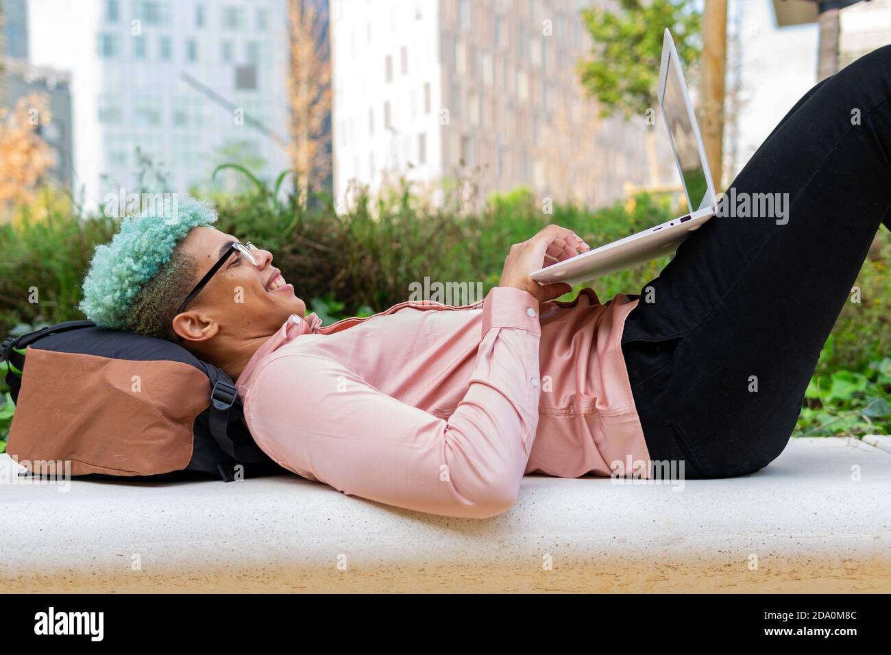 Side view of smiling African American male student with blue hair lying on backpack in university campus and typing on laptop while working on homewor Stock Photo