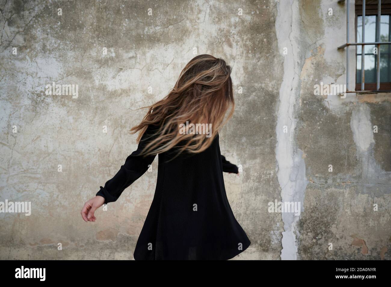 Unrecognizable female in black dress and long hair covering face standing against grungy concrete wall of aged building Stock Photo