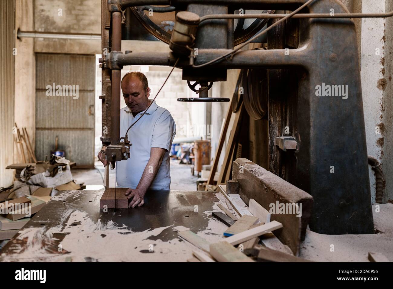 Serious male carpenter using band saw and cutting wooden plank at workbench in messy workshop Stock Photo