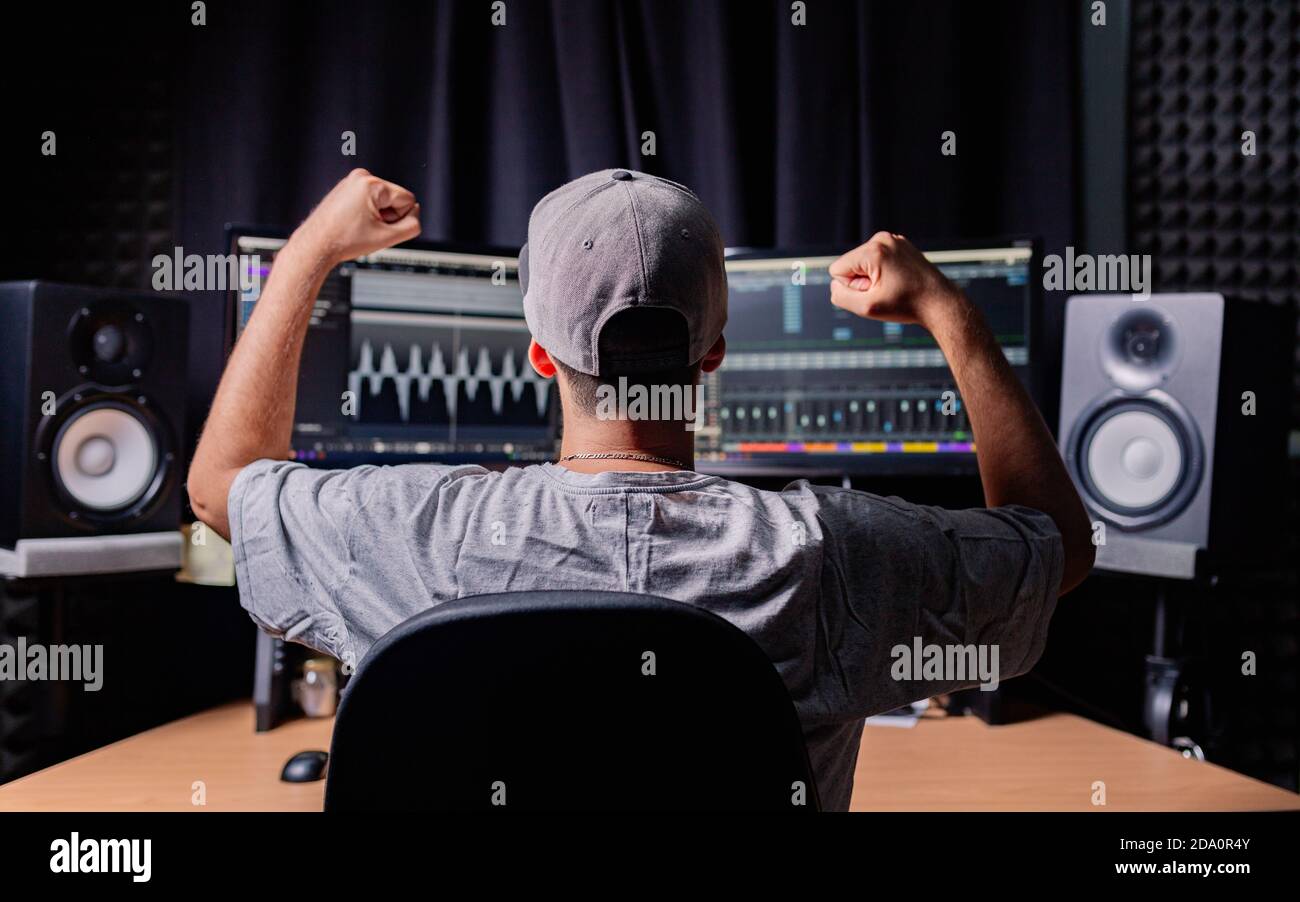 Back view of unrecognizable male musician sitting at table with monitors and stereo speakers while celebrating victory with fists up and recording aud Stock Photo