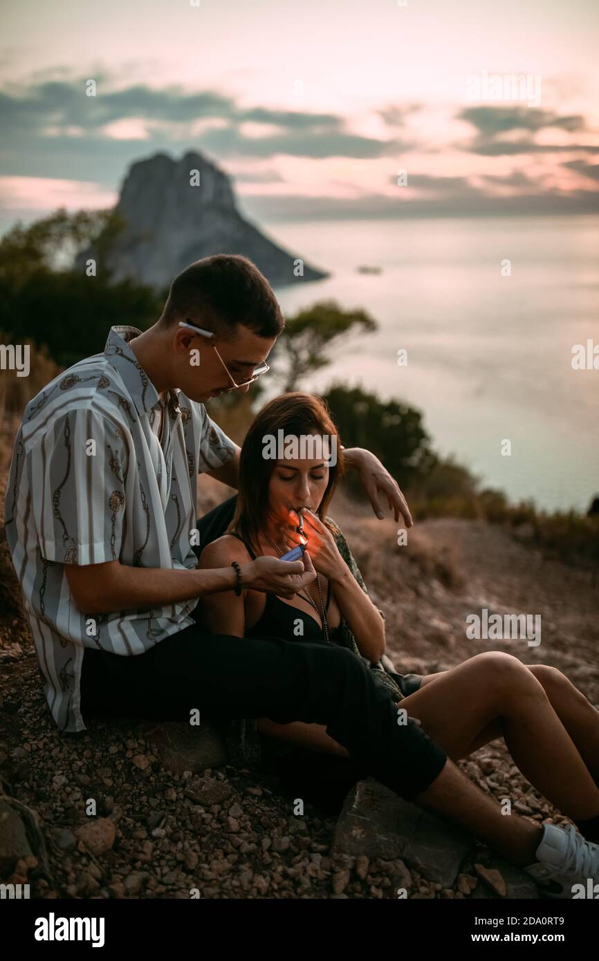 cigarette with girlfriend Side view of calm man lighting cigarette for girlfriend while chilling on  seashore at sunset Stock Photo
