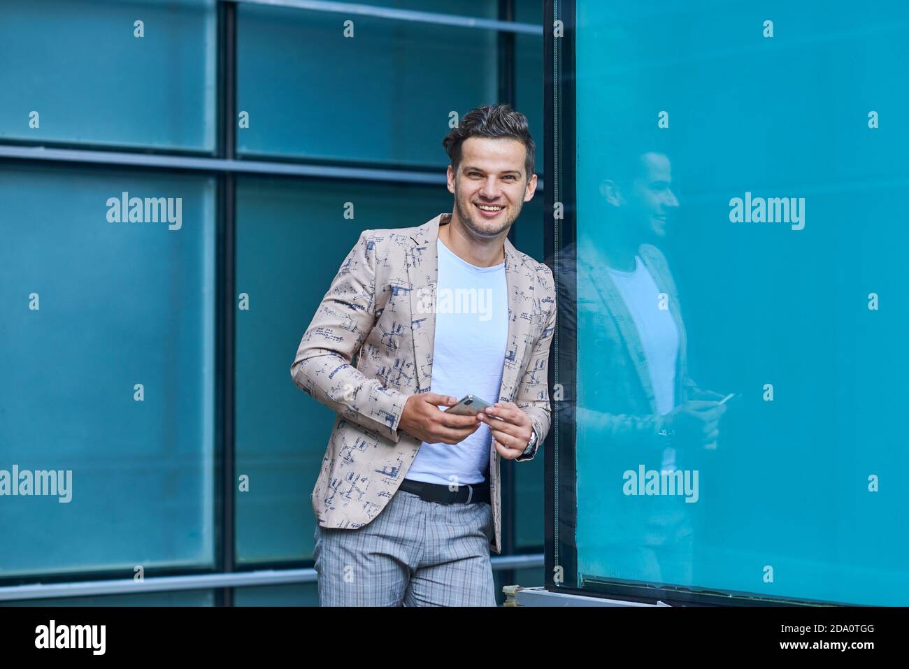 Young glad office worker in trendy clothes with cellphone looking at camera while leaning on wall in city Stock Photo