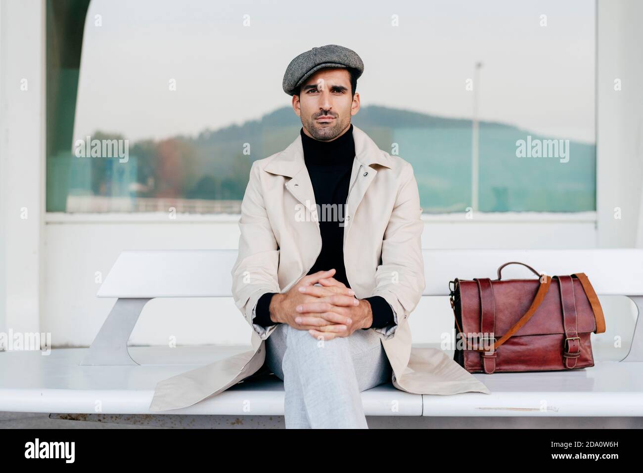 Confident man with beard in trendy clothes sitting on white bench with bag and looking at camera against modern building with big window Stock Photo
