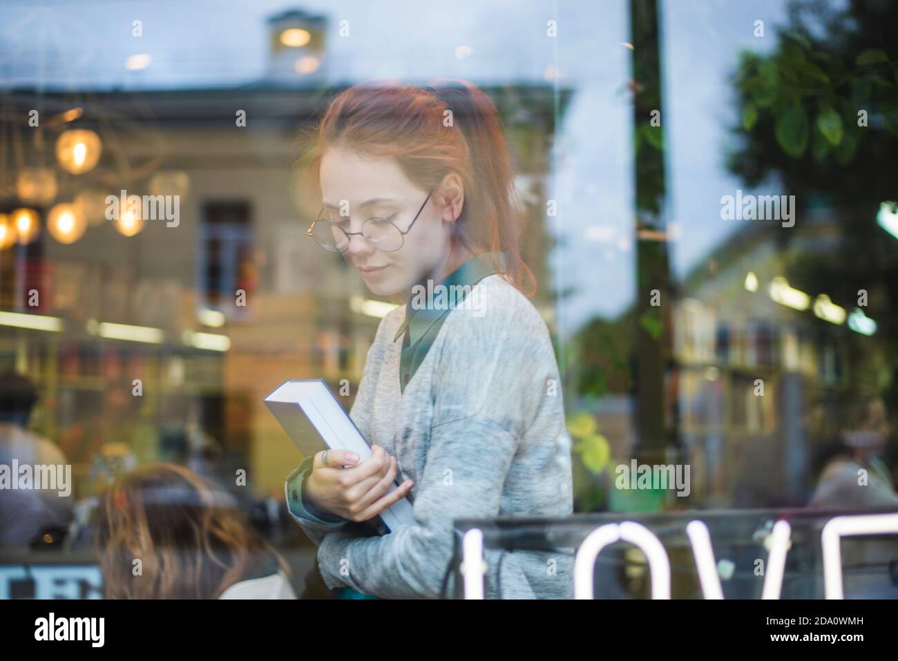 Through window side view of young stylish female in eyeglasses sitting in cozy bookshop with volume Stock Photo