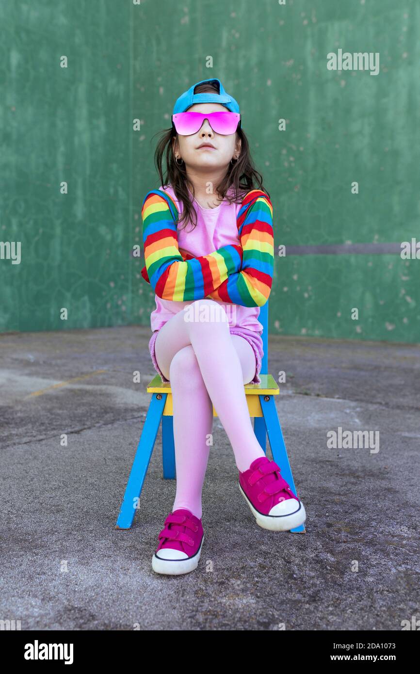 Confident girl wearing trendy outfit and pink sunglasses sitting with crossed arms and legs on chair and looking at camera Stock Photo