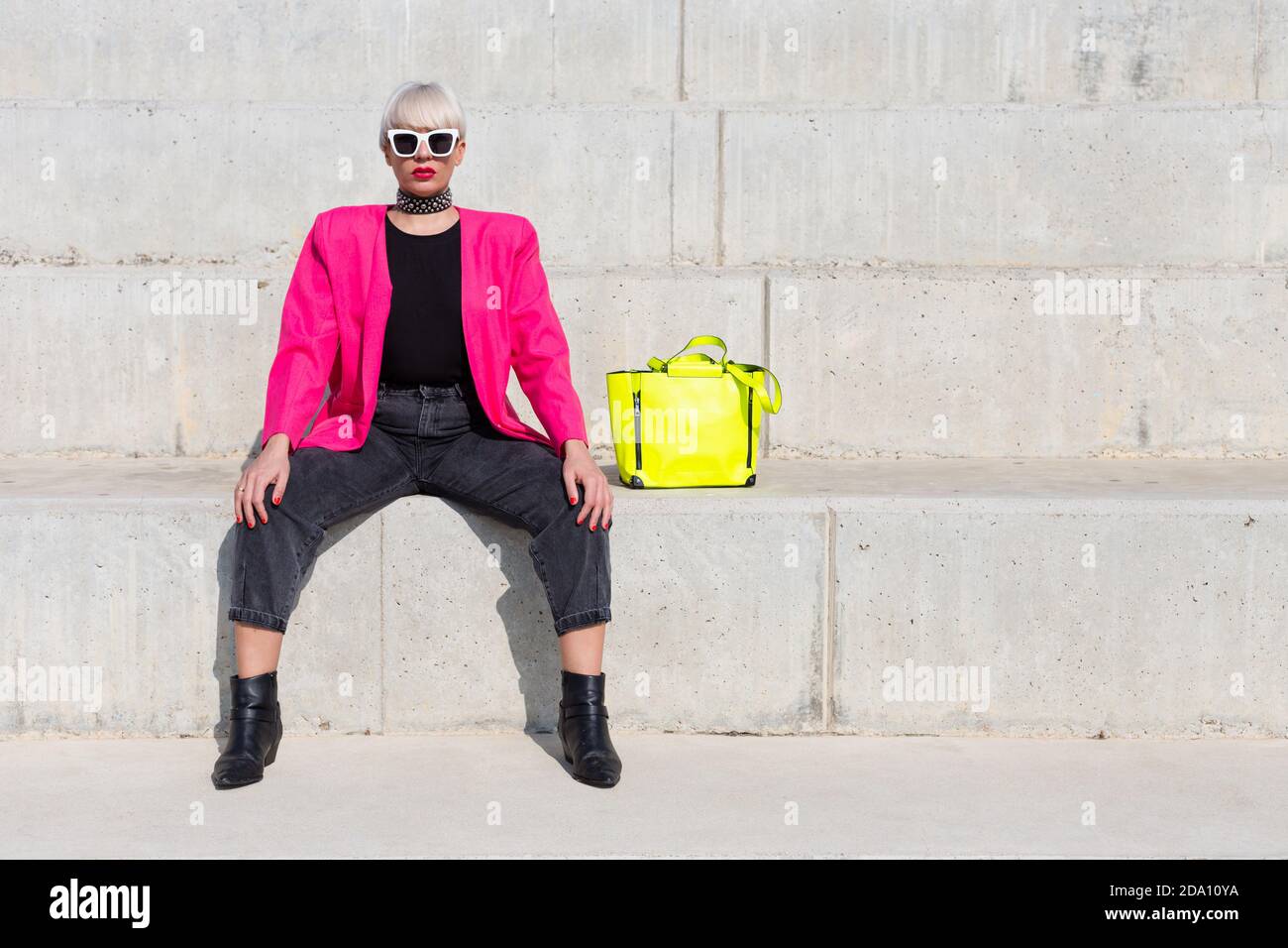 Confident female model wearing bright pink jacket sitting on stairs with vivid yellow handbag in city and looking at camera Stock Photo