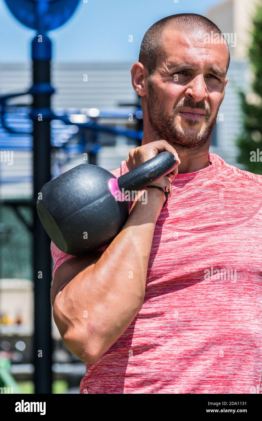 Serious male athlete with strong body doing exercises with heavy metal kettlebell during intense workout in summer Stock Photo