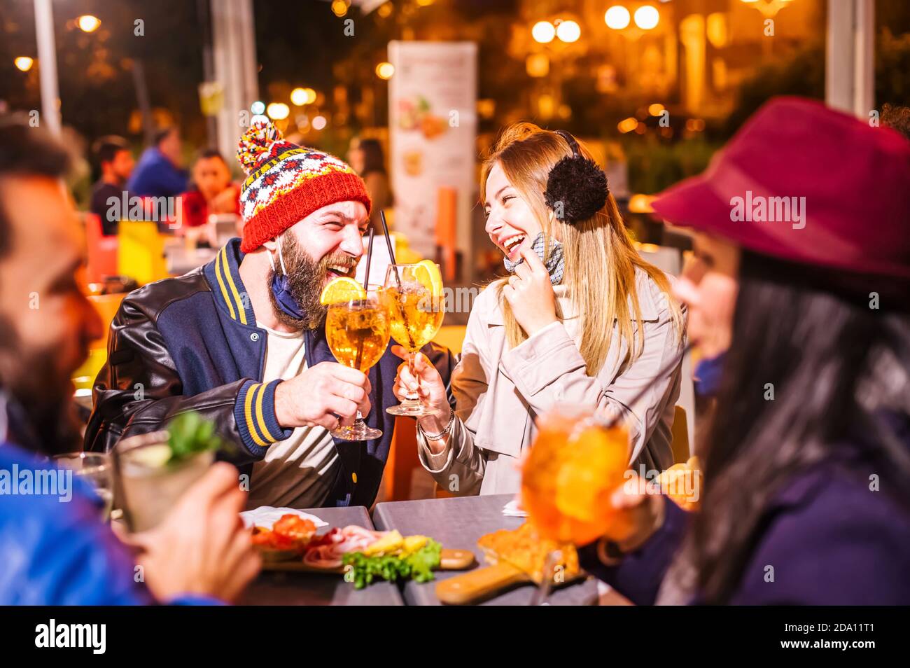 Man and woman smiling and toasting spritz at winter cocktail bar outdoors wearing open face mask - New normal lifestyle concept with millennial people Stock Photo