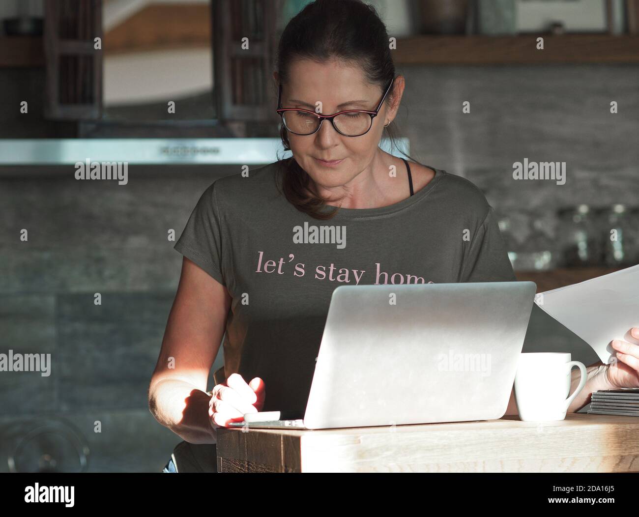 Middle aged caucasian woman with glasses working at home - in the kitchen - with laptop and documents. Stay home during covid-19 quarantine concept.  Stock Photo