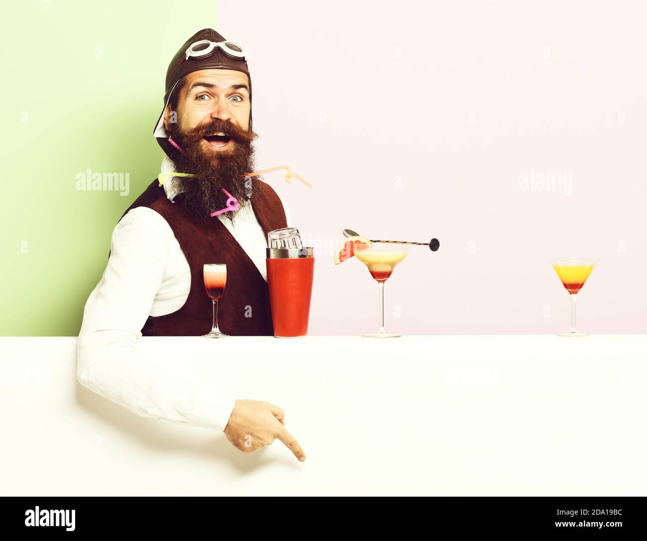 handsome bearded aviator or pilot with long beard and mustache on smiling face with shaker alcoholic cocktail in vintage suede leather waistcoat with hand and glasses on purple green studio background Stock Photo