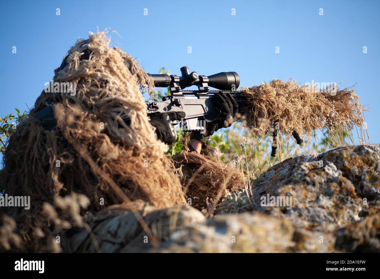 sniper in ghillie (camouflage) suit with a high-precision rifle with optic scope in a combat position in the mountains Stock Photo