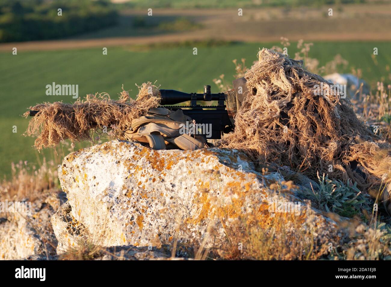 sniper in ghillie (camouflage) suit with a high-precision rifle with optic scope in a combat position in the mountains Stock Photo