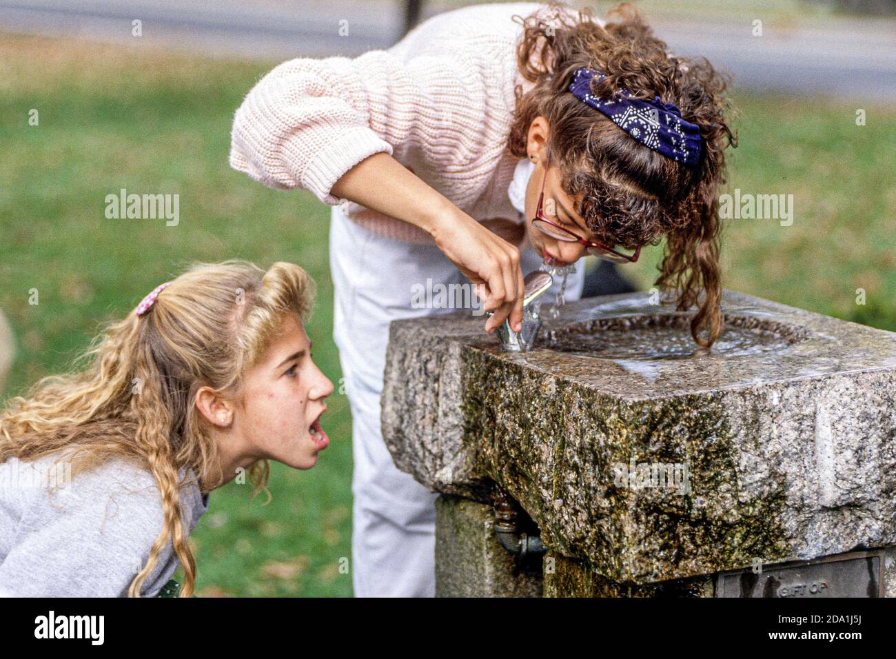 A young girl drinking from a water fountain with her friend looking on Stock Photo Alamy