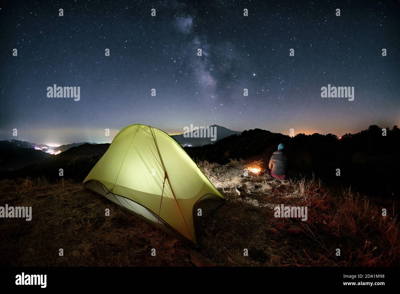 hiker overlooking milky way by the fire nearby illuminated tent in Malabotta Nature Reserve, Sicily Stock Photo