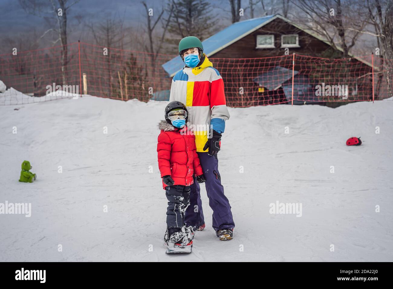 Snowboard instructor teaches a boy to snowboarding wearing a medical mask during COVID-19 coronavirus. Activities for children in winter. Children's Stock Photo