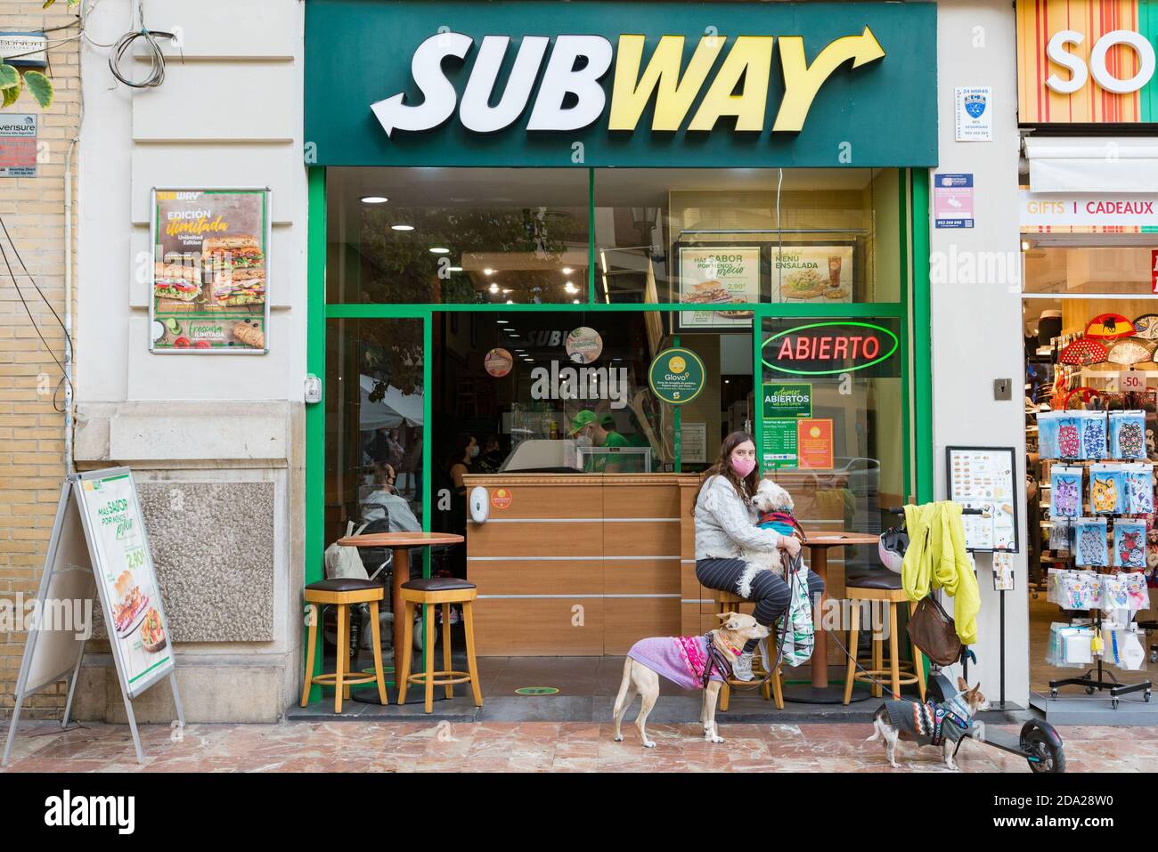 A woman wearing a face mask sits outside the Subway fast food restaurant with her three dogs. Stock Photo
