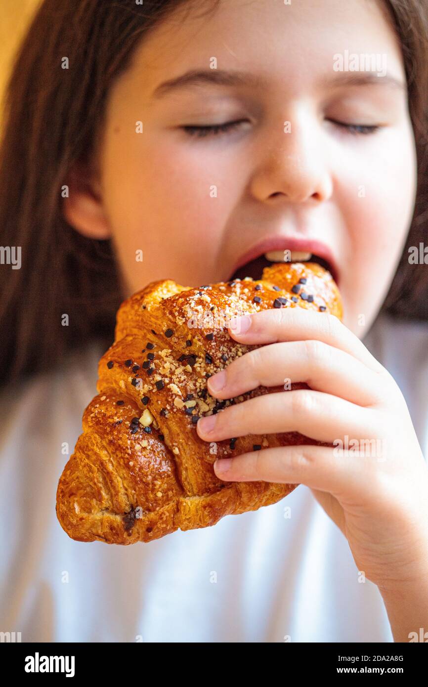Brunette girl in a white T-shirt eating a chocolate croissant, orange background. Child eating dessert concept. Stock Photo