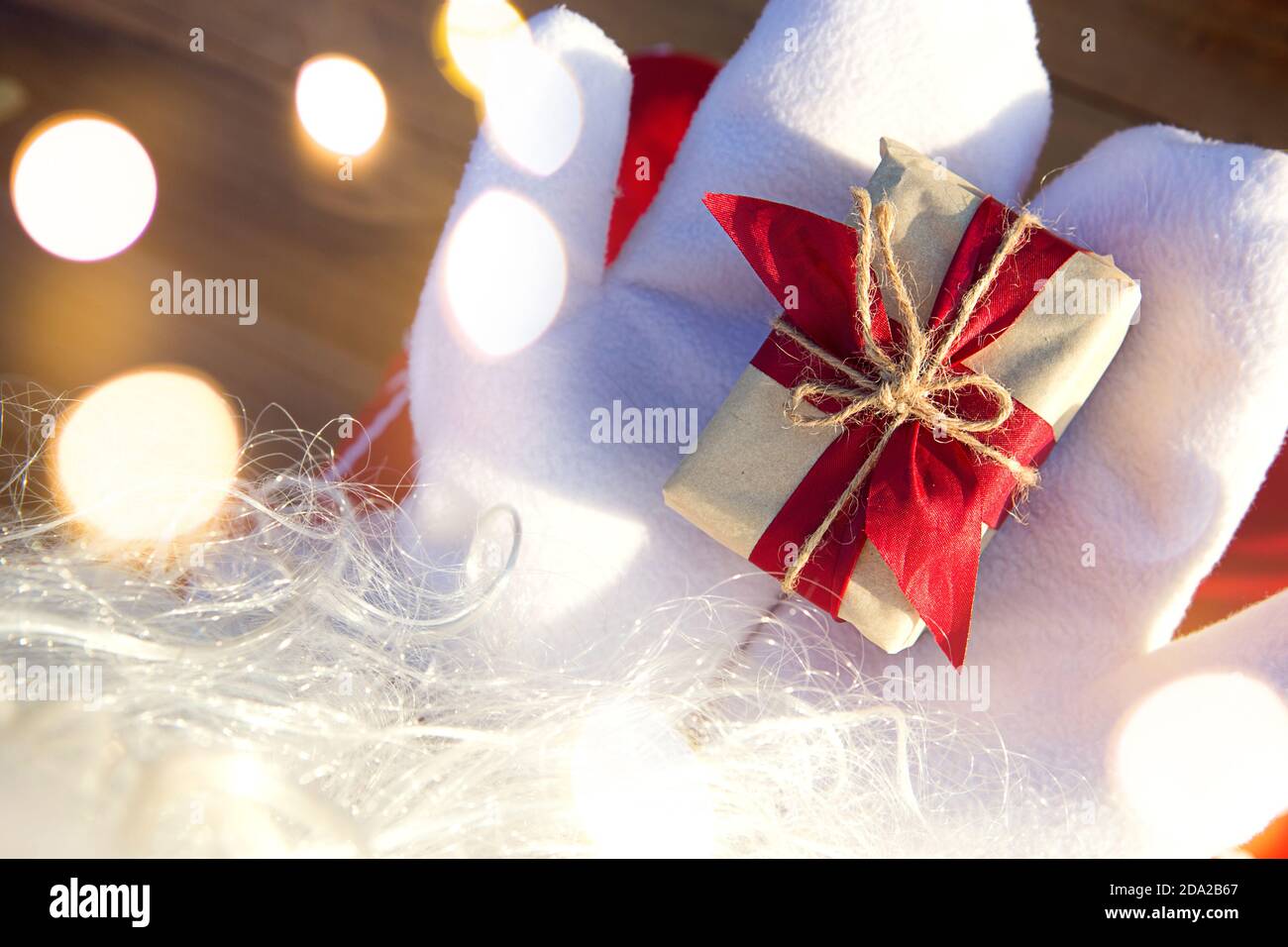 A box with a Christmas gift in the hands of Santa Claus in white mittens. Red suit, beard, garland lights in a blur. New year, preparation, expectatio Stock Photo