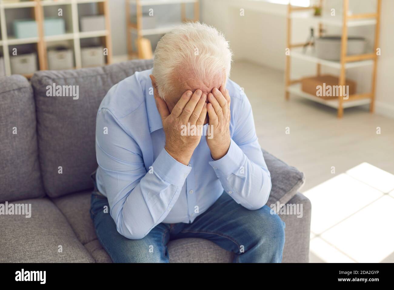 Shocked senior man covers his face with his hands from grief sitting at home on the couch. Stock Photo