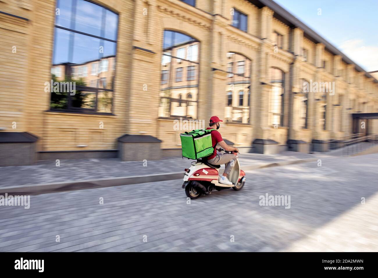 Fast delivery during covid 19 outbreak. Rear view of a male courier with thermal backpack wearing black protective mask riding scooter along the street, selective focus on a man. Stay home concept Stock Photo