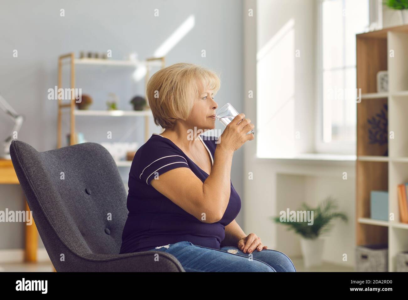 Mature woman sitting in armchair and drinking pure water at home Stock Photo