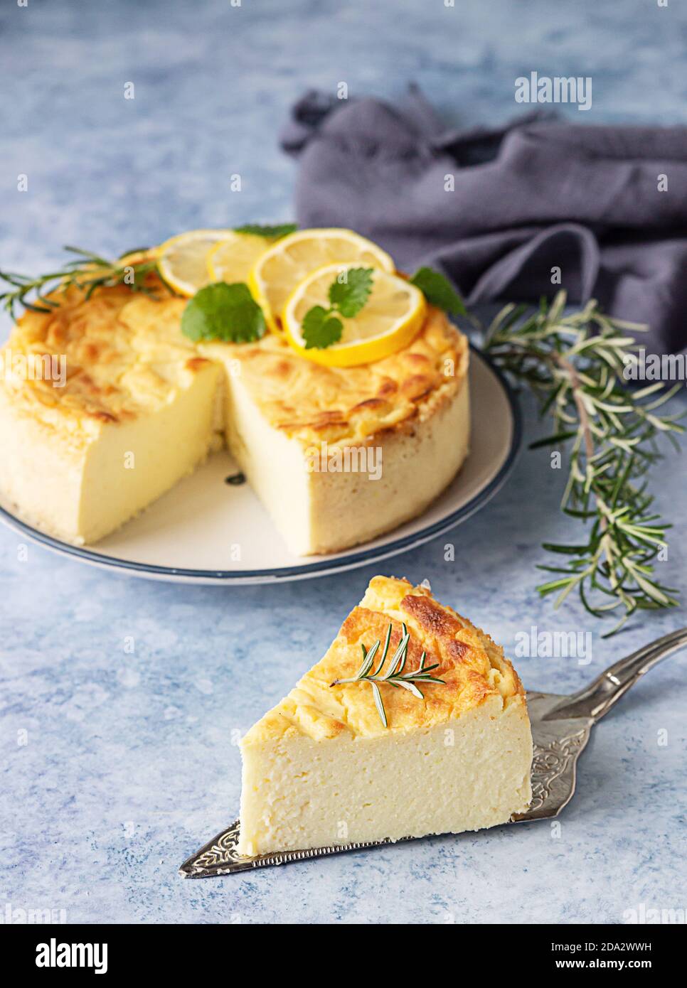 Lemon vanilla cheesecake decorated with rosemary, mint and lemon slices. Ricotta no crust cheesecake or casserole. Blue concrete background. Stock Photo