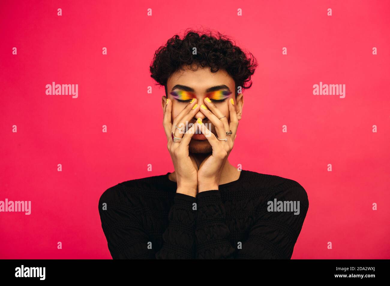 Gay man wearing rainbow colored eye shadow. Androgynous male holding his face with eyes closed on red background. Stock Photo